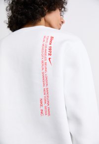 Sweatshirt branco com texto vermelho nas costas, listando cidades e "Desde 1972." Feito de tecido, possui gola redonda e um corte descontraído.