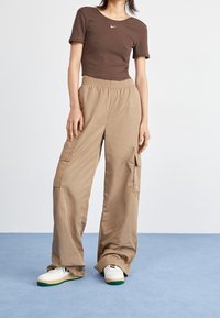 Braun eng anliegendes T-Shirt mit kleinem weißem Logo, kombiniert mit beigen Cargo-Hosen mit Seitentaschen und lockerer Passform, getragen mit weißen Sneakers.