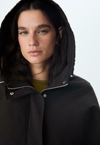 Čierna kapucňová bunda so zipsom a zatváraním na cvoky, s hladkou textúrou a jemným leskom, nosená na olivovo zelenej blúzke.