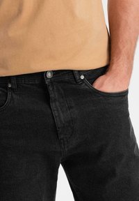 Pantalones vaqueros de mezclilla negra con un corte clásico, que presentan una cremallera, cierre de botón y bolsillos frontales. La tela tiene una textura lisa y mate.