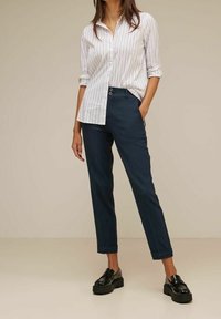 Bílá pruhovaná košile s knoflíky a ohrnutými rukávy, spárovaná s tmavě modrými zúženými kalhotami a černými mokasíny s robustní podrážkou.