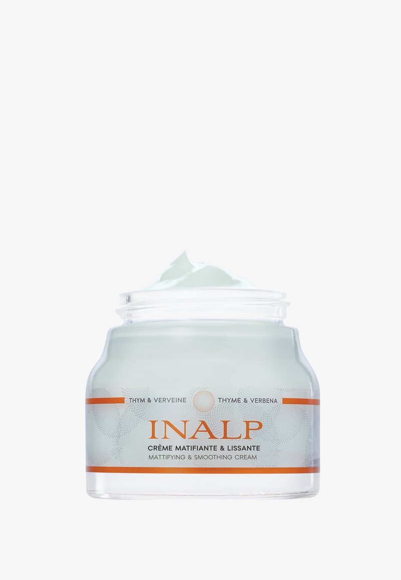 Bocal en verre clair de forme arrondie, contenant une crème vert clair. Présente un couvercle blanc et une étiquette orange avec du texte et de petits motifs circulaires.