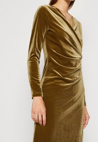 Robe en velours vert olive à manches longues, avec un décolleté en V et des détails froncés à la taille, créant une apparence texturée.