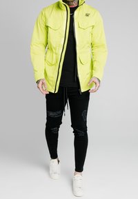 Neon gul jakke med to forlommer, lynlås lukning og høj krave, parret med sorte slidte jeans og hvide sneakers.