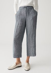 Une personne portant un pantalon en lin gris à taille haute et jambes larges avec des plis sur le devant et des mocassins blancs, les mains dans les poches, sur un fond uni.