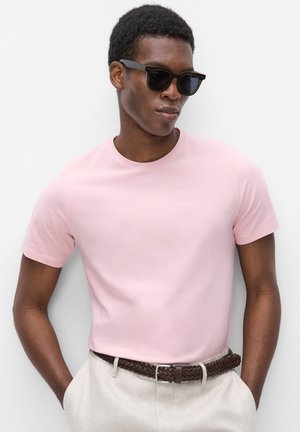 Man draagt zwarte zonnebril, lichtroze T-shirt met korte mouwen, beige broek en een donkerbruine gevlochten riem, staand met handen in de zakken.
