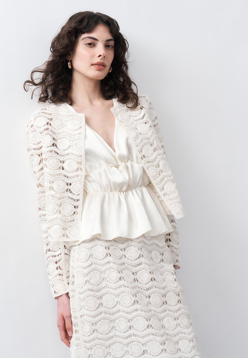 Γυναίκα με σγουρά σκουρόχρωμα μαλλιά φορά λευκό δαντελένιο σακάκι και φούστα πάνω από λευκό σατέν τοπ με πεπλούμ, στέκεται μπροστά σε απλό φωτεινό φόντο.