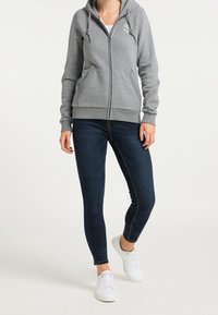 Felpa grigia con zip e cappuccio, dotata di cordini e tasche frontali, abbinata a jeans skinny blu scuro e sneakers bianche. Design semplice e texture casual.