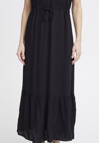 Robe maxi noir sans manches avec un ourlet à volants, taille ajustable à nouer, et texture de tissu douce, offrant une coupe décontractée et une silhouette fluide.