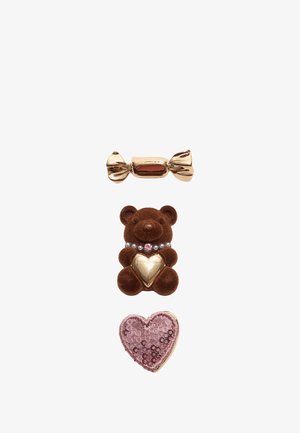 Drei dekorative Broschen: eine in Gold gewickelte Süßigkeit, ein brauner Teddybär mit einem Herzen und ein pinkfarbener, mit Pailletten besetzter Herz, vertikal angeordnet.