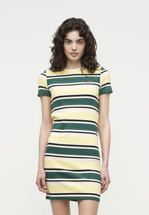 Kvinne med krøllete mørkt hår, iført en kortermet kjole som rekker til knærne med horisontale striper i grønt, gult, hvitt og svart.