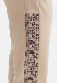 Pantaloni jogger beige con una pannello laterale che presenta un motivo con logo ripetuto in tonalità scure e chiare. Tessuto morbido e design aderente.