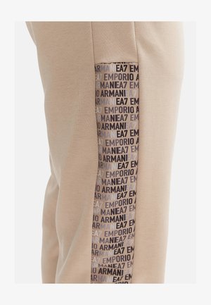 Pantaloni jogger beige con una pannello laterale che presenta un motivo con logo ripetuto in tonalità scure e chiare. Tessuto morbido e design aderente.