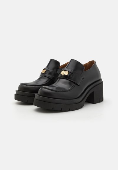 Schwarze Lederschuhe mit einer klobigen Plateausohle, die mit einem dekorativen Riemen mit einem goldenen Emblem und gesteppten Details an der Vorderseite versehen sind.