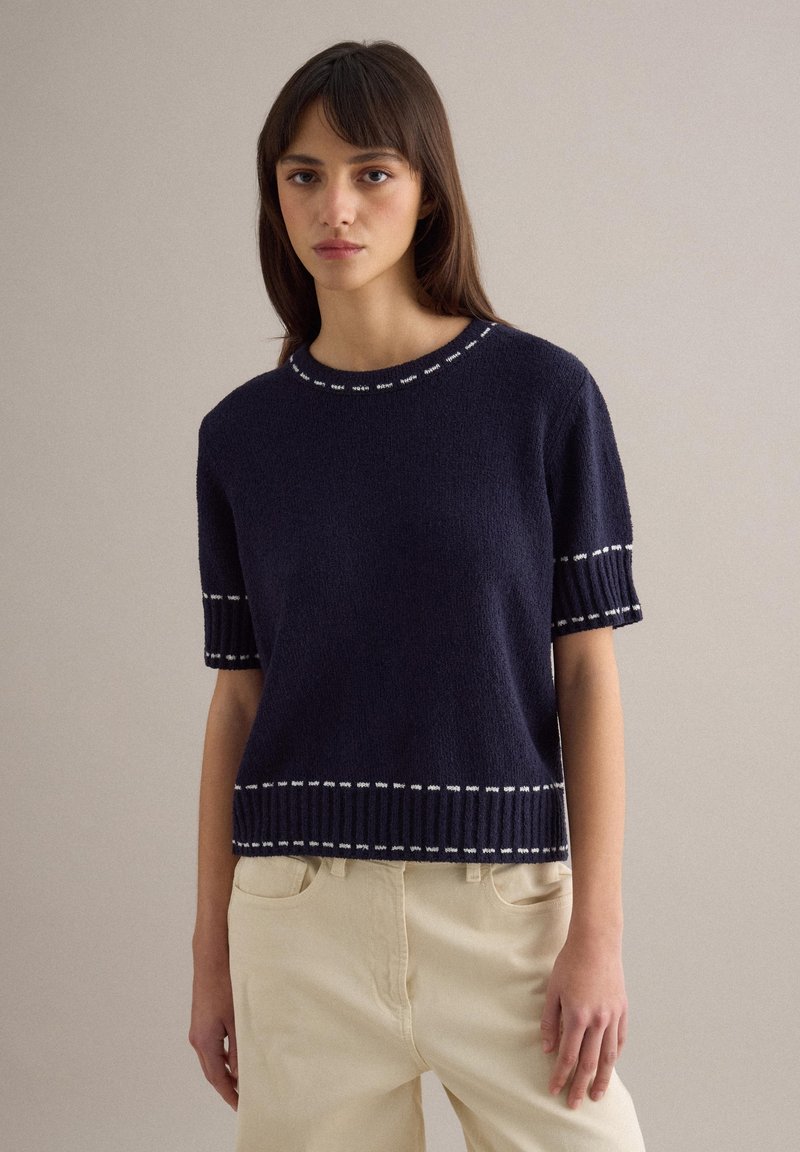 Joven mujer con un suéter de punto azul marino de manga corta con ribete blanco, combinado con pantalones beige de talle alto, frente a un fondo liso.