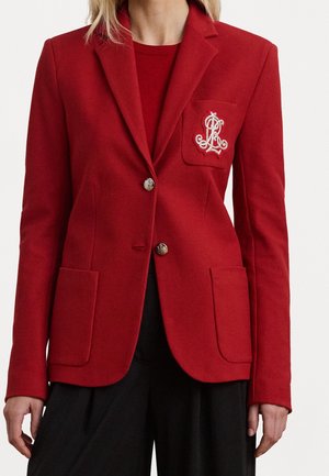 Kvinde iført en rød blazer med guldknapper og hvide broderede initialer på brystlommen, kombineret med sorte bukser.