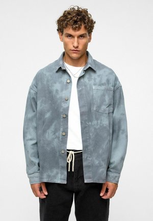 Vervaagde blauw-grijze oversized shirt met een knoopsluiting aan de voorkant, een borstzak en lange mouwen, gecombineerd met een wit t-shirt en zwarte broek.