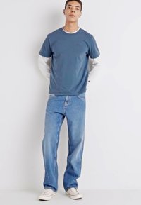 De mannenoutfit bestaat uit een blauwe T-shirt met korte mouwen over een witte overhemd met lange mouwen, gecombineerd met lichtblauwe baggy jeans en witte sneakers.