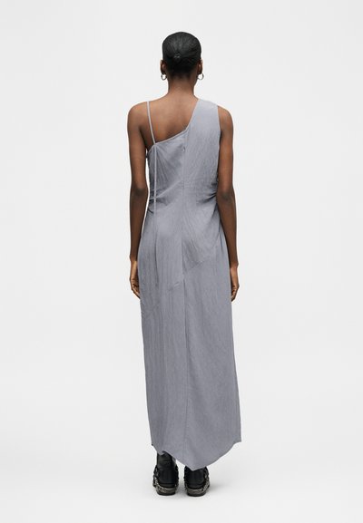 Γυναίκα με σκουρόχρωμο δέρμα και μαλλιά σε κότσο, ντυμένη με μακρύ, αμάνικο, ασύμμετρο γκρι φόρεμα και μαύρα αθλητικά παπούτσια, στέκεται με την πλάτη στην κάμερα.