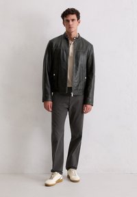 Junger Mann steht in einer schwarzen Lederjacke, einem beigen Henley-Shirt, dunkelbeigen Hosen und weißen Sneakers vor einer schlichten Wand.