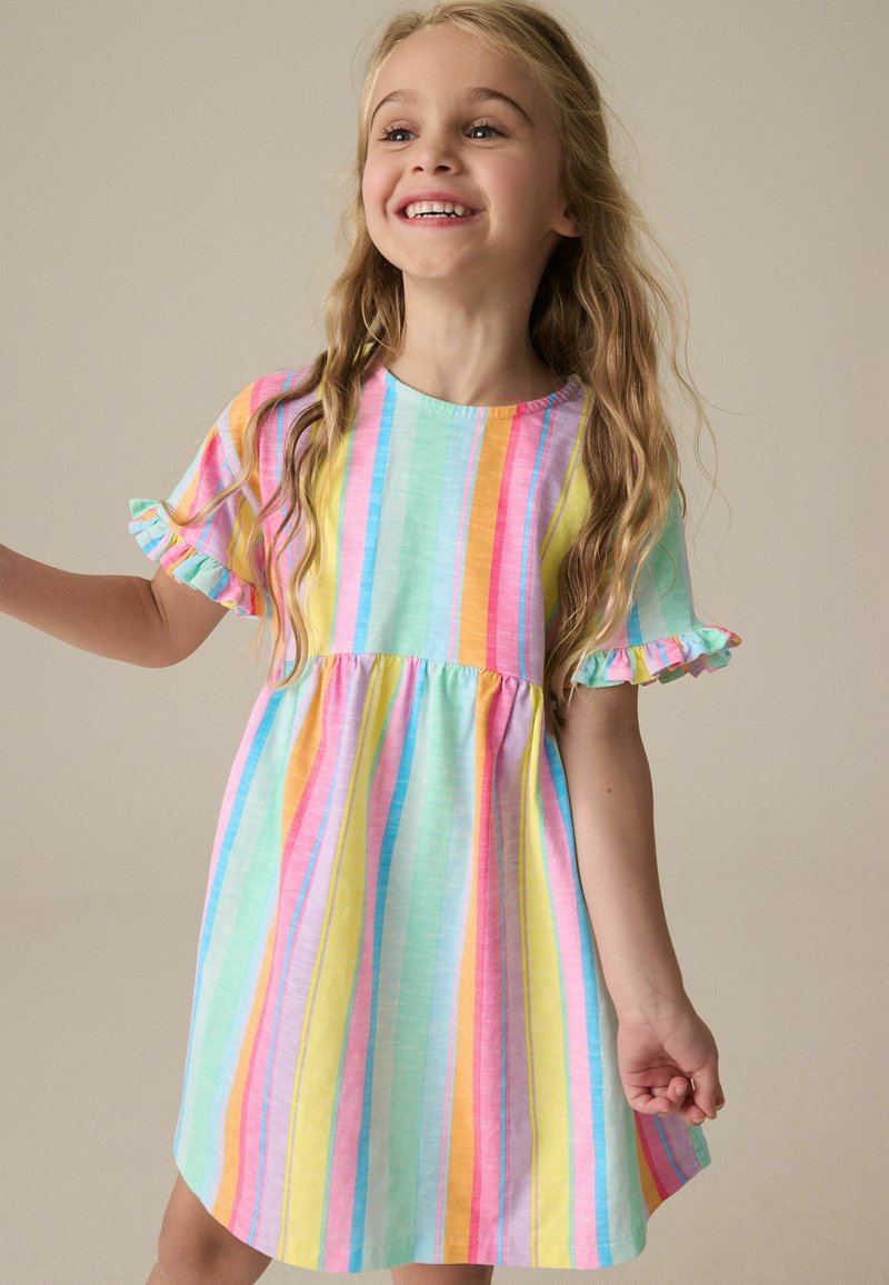 Ragazza giovane sorridente, indossa un vestito a righe arcobaleno pastello con maniche corte arricciate, in piedi davanti a uno sfondo beige uniforme.