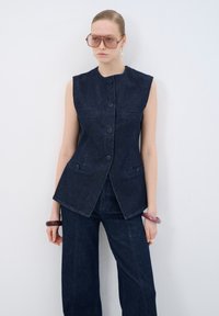 Chaleco de denim en azul oscuro con un cuello alto, diseño sin mangas, botones frontales, dos bolsillos en el pecho y un dobladillo ligeramente acampanado, combinado con pantalones a juego.