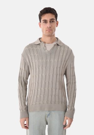 Uomo che indossa un maglione beige lavorato a trecce con colletto sopra una camicia chiara e jeans azzurri, in piedi su sfondo bianco.
