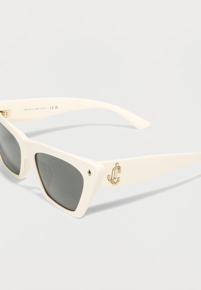 Λευκά γυαλιά ηλίου cat-eye με σκούρους φακούς, διακοσμημένα με ένα μικρό χρυσό λογότυπο και διαμαντένιο σχέδιο στη γωνία του μπροστινού μέρους του σκελετού.