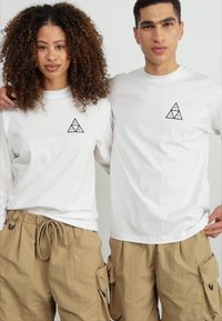 Langermede hvite skjorter med svart trekantet logo på brystet, paret med beige cargo-shorts med sidelommer og snøring i livet.