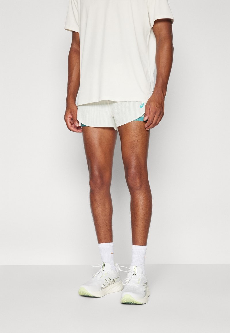 Homme portant des chaussures de course blanches, des chaussettes blanches, un short de sport blanc avec une bordure turquoise et un t-shirt blanc ample.