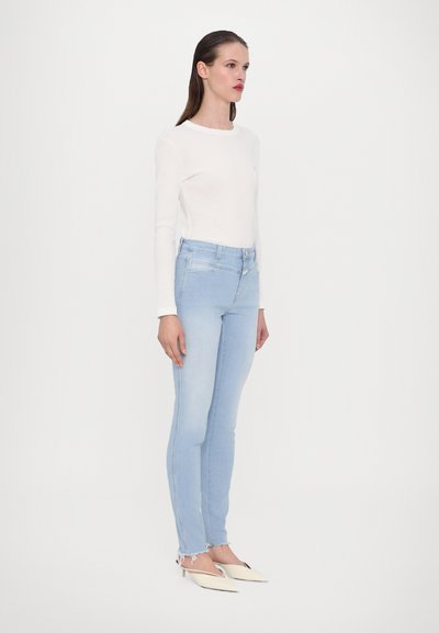 Hvit langermet skjorte, lyseblå skinny jeans med frynsete kant, beige slingback-sko, stående mot en enkel hvit bakgrunn.