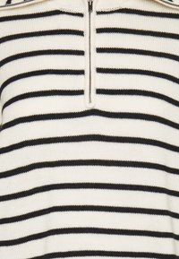 Gros plan sur un pull en maille blanche avec des rayures horizontales noires et une fermeture éclair partiellement fermée au col.