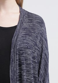 Cardigan a righe blu navy e bianche con apertura frontale e tessuto morbido e testurizzato. Leggero e cade in modo fluido su una maglietta nera.
