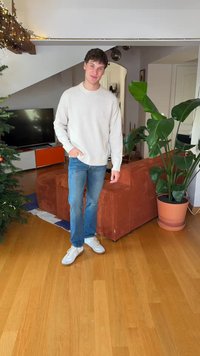 Svetlo siva pletena majica, modre kavbojke in bele superge. Rjava kordurova kavč z rastlino v bližini. Lesena tla z vidnim preprogo.