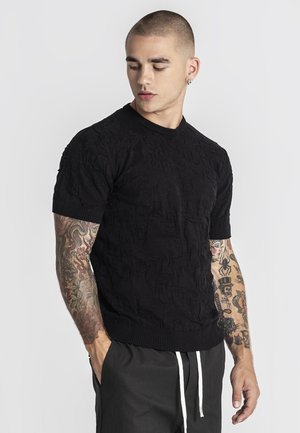 Jonge man met kort haar en volledige mouwtattoos draagt een gestructureerd zwart shirt met korte mouwen en zwarte broek met witte trekkoorden.