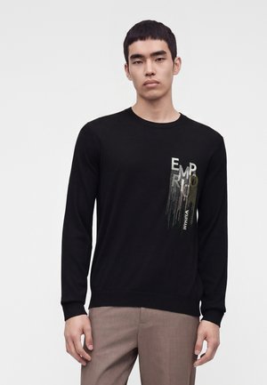 Jeune homme aux cheveux noirs courts portant une chemise noire à manches longues avec un motif de texte vertical "EMPORIO ARMANI" et un pantalon beige.
