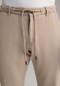 Gros plan d’un pantalon beige avec un cordon en tissu passant par les passants de ceinture, une fermeture par bouton et de légères rides sur le tissu.