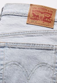 Jeans de mezclilla azul claro con una etiqueta de cuero que presenta gráficos y texto en rojo. Detalles de costura destacados y un diseño clásico de bolsillo curvado.