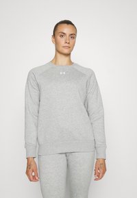 Sudadera gris con cuello redondo y mangas raglán. Fabricada en tejido suave, con un pequeño logotipo en el pecho. Combinada con pantalones grises a juego.