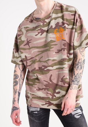 Person trägt ein lockeres Camouflage-T-Shirt mit orangenen Buchstaben auf der Brust und schwarze zerrissene Jeans, zeigt Tattoos auf beiden Armen und Händen.