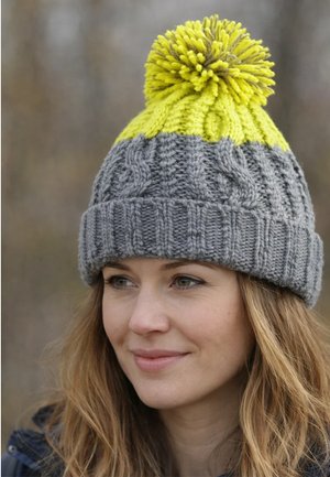 Woman with light brown hair wears a gray knit hat with yellow top and pom-pom, looking slightly right outdoors in soft natural light.
