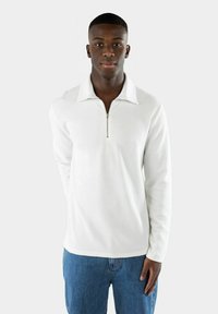Vit långärmad tröja med dragkedja i kragen, ribbad textur och rak fåll, tillsammans med blå jeans.