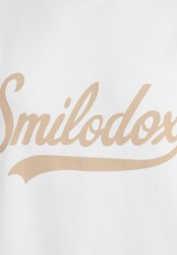 Hvidt stof med et stort, kursiv beige logo, der står "Smilodox", centreret og i en fremtrædende position.