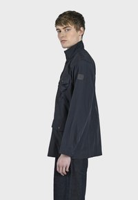 Černá lehká bunda s vysokým límcem, předními kapsami a knoflíkovými detaily. Vyrobena z odolné látky, má na rukávu nášivku s logem.