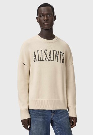 Mann trägt einen cremefarbenen Pullover mit dem Schriftzug "ALLSAINTS" auf der Brust und blaue Jeans, steht vor einem schlichten hellen Hintergrund.