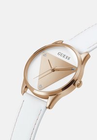 Kuldtooneline kolmnurkne kellanägu valge taustaga, kuldsed käed, GUESSi logo ja valge õmmeldud nahkrihm.