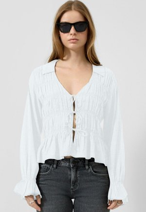 Kobieta w czarnych okularach przeciwsłonecznych, białej bluzce z marszczeniami i zawiązywanymi wiązaniami oraz czarnych jeansach z wysokim stanem, stojąca na jednolitym tle.