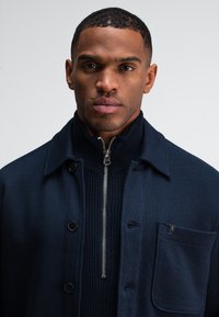 Navyblauwe jas met een ritskraag over een gebreide trui. Voorzien van een knoopsluiting, een borstzak en zilverkleurige hardware accenten.