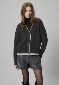 Cárdigan gris oscuro con ribetes crema, cierre de botones y corte relajado. Combinado con pantalones cortos oscuros y un cuello de tortuga negro para superponer.