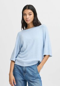 Světle modrý pletený svetr s širokým výstřihem, krátkými rukávy a žebrovaným lemem. Měkká textura a volný střih, kombinovaný s modrými džíny.